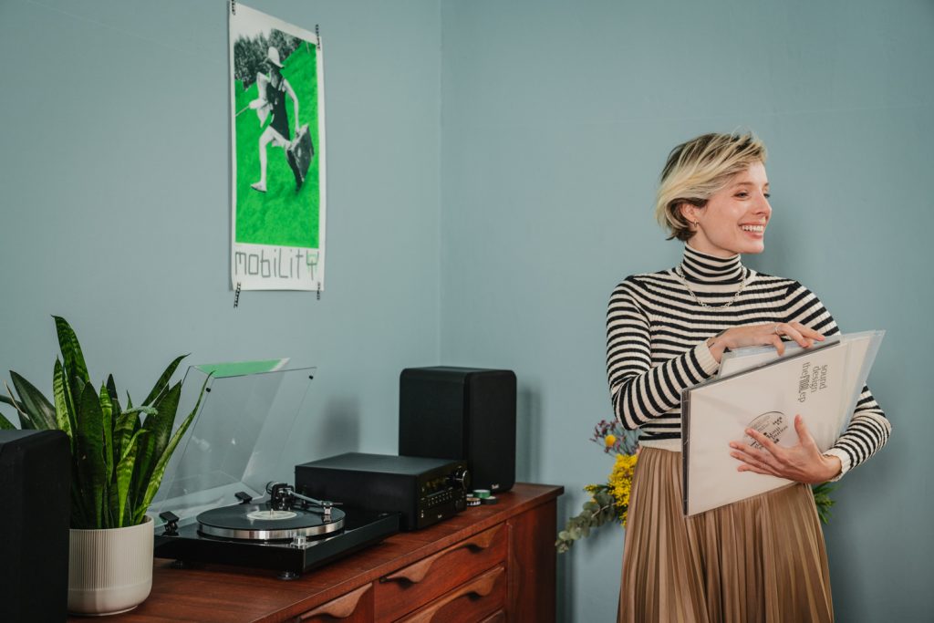 femme souris avec des pochettes de vinyles dans la main derrière elle un meuble avec un tourne-disque et les enceintes Teufel 
