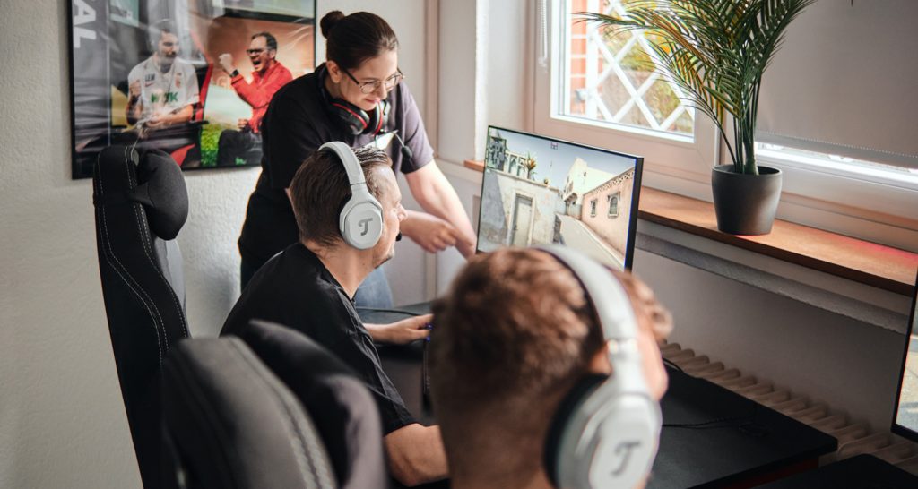 un homme joue aux jeux vidéos sur pc avec un casque gaming Teufel, son amie lui montre quelque chose sur l'écran et un autre ami joue aussi sur PC aux jeux vidéos avec un casque audio gaming Teufel CAGE ONE